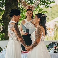 Das Bild zeigt zwei Bräute in weißen Hochzeitskleidern, die sich lächelnd an den Händen halten, während eine Hochzeitsbeamtin