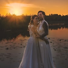 Das Bild zeigt zwei Bräute in weißen Hochzeitskleidern, die sich bei Sonnenuntergang auf einem Holzsteg an einem See umarmen.