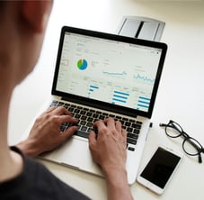 Homem analisando gráficos em um laptop, com óculos e smartphone sobre a mesa.