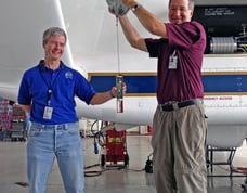 Two persons holding a dropsonde and its parachute beside Global Hawk drone for science and weather.