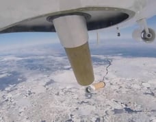 Under belly of plane image of dropsonde departing aircraft above a snow covered landscape far below.