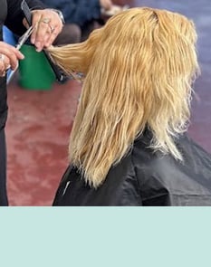 Blonde woman receiving a hair trim, seated in a chair with a stylist behind her.
