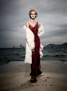 Model wearing evening dress and coat standing on a dock with the ocean and ships in the background