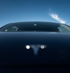 Low‑angle front view of an electric car’s hood TESLA and windshield under a blue sky, By ACAT Photos