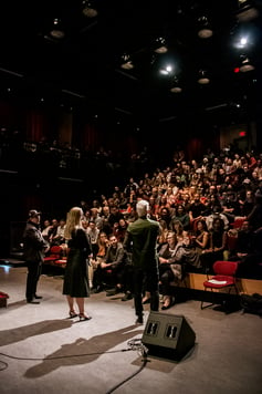 A host stands in the spotlight, mic in hand, on a theatre stage in front of a large audience