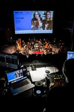 A technician operates a console while an orchestra plays on the stage below in front of a screen