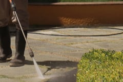 person in wellies uses pressure washing wand to clean stone floor near an hedge