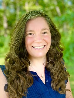 Shannon Gale, with long brunette hair, and a navy shirt, smiling in front of a green woodland scene