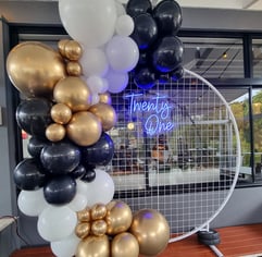 White circle mesh backdrop with blue Twenty One neon sign and a gold, black and white balloon garland, Redlands
