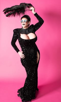 regal drag queen in long sequined black gown holding a large feather fan overhead on pink backdrop