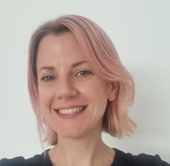A head and shoulders picture of woman with pink hair, smiling, wearing a black top