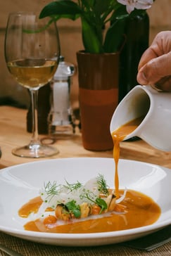 Sopa de pescado vertida en mesa con marisco, hinojo y hierbas frescas, junto a una copa de vino blanco