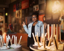 A Black woman in a black dress and blue jacket standing among a wooden stools upturned on tables