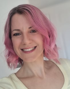 A head and shoulders image of a woman with pink hair, smiling, wearing a yellow top.