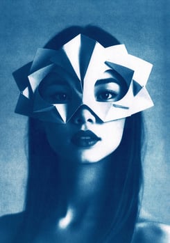 Close-up of a woman with long, straight hair, wearing a geometric paper mask covering her upper face
