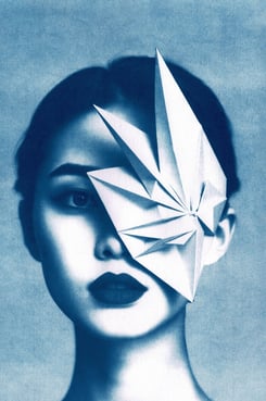 Close-up of a woman with blue-toned skin, dark lips, and a geometric paper structure covering her ri