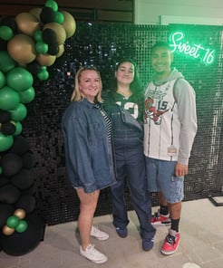 Ashlee with her daughter and husband posing in front of the black shimmer wall and green Sweet 16 neon sign Brisbane