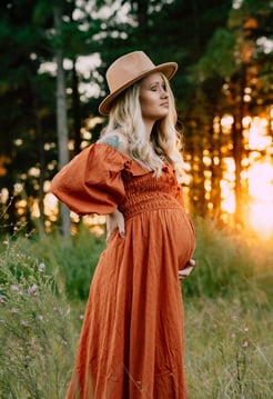 a pregnant blonde woman in a dress and hat, standing in a field