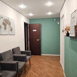 Sala de espera com cadeiras cinza, quadros na parede, portas com sinalizações e prateleira com enfeites.