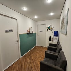 Sala de espera com cadeiras cinza, mesa com garrafas de água e café, plantas decorativas, sinalizações nas portas e quadros.