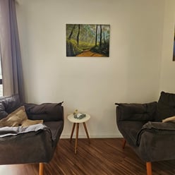 Canto de uma sala com duas poltronas cinza escuro, mesa branca redonda com item decorativo, quadros pendurados nas paredes.