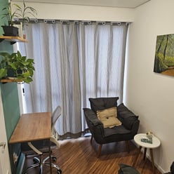 Consultório com cadeira de escritório, mesa de madeira, poltronas cinzas com almofadas, cortina e plantas.