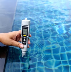Hand holding a digital salt meter to test pool water quality with reflection of blue tiles.