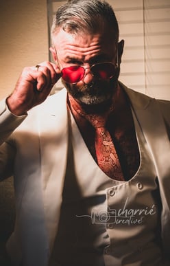 dapper bearded man dressed in formal clothing in low light staring intently over his glasses