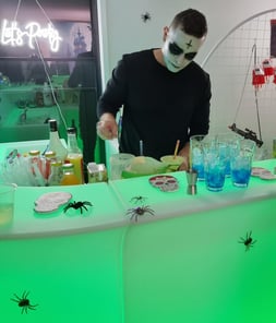 Bartender in Halloween makeup preparing drinks at a glowing green LED bar decorated with crawling spiders.