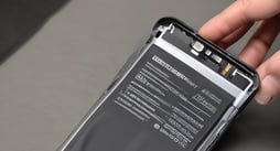 Two hands are exchanging a smartphone over a glass counter. The screen of the phone is cracked, indicating possible damage or need for repair. One person is wearing a brown shirt, and the counter displays other phones and electronic devices.