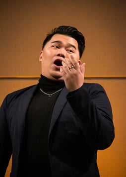 A male Asian opera singer in formal clothing emphasizes his singing with a hand gesture