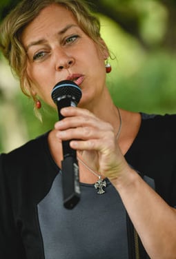 Eine blonde Frau mit grünlichen Augen spricht in ein Mikrofon, wobei ihr Blick leicht nach oben gerichtet ist.