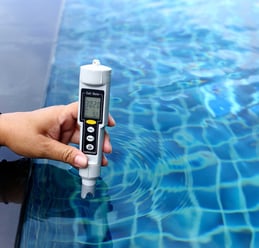 Hand holding a digital salt meter to test pool water quality with reflection of blue tiles.
