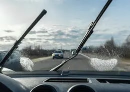 View from inside a car as windshield wipers clear washer fluid while driving on a sunny day with light traffic ahead.