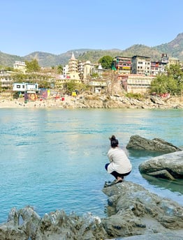 Eva standing by the sacred Ganges River in Rishikesh, India, with mountains visible in the background on a sunny day.