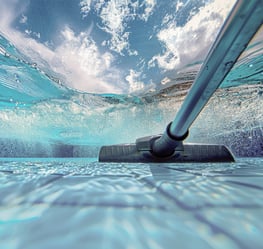 Technician skimming a residential pool with a long cleaning pole under bright sunlight.