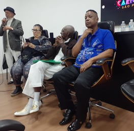 Foto tirada do simpósio, na foto três pessoas sentadas em cadeiras. Um homem ao fundo falando no mic