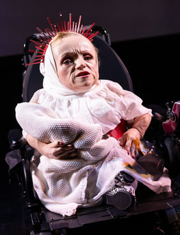 A white woman of very short stature in an electric wheelchair wearing white, with a crown of spikes