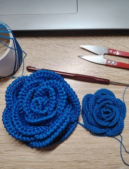 rose faite au crochet dans un fil fin de couleur bleu posé sur une table un crochet et un ciseaux
