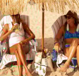 two women sitting on a beach under a straw umbrella