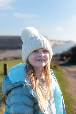 A profile photo of Poppy Walker, smiling wearing a beanie hat and a puffer jacket against the backdrop of Cuckmere Haven