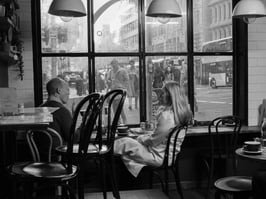 A man and woman sitting in a coffee shop next to a broad window through which many people can be seen.