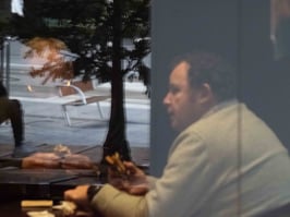 Two men sitting at coffee shop table. One is hidden by window reflection.