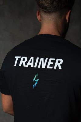 Spatele unui antrenor personal, purtând un tricou negru inscripționat cu „TRAINER” și un logo