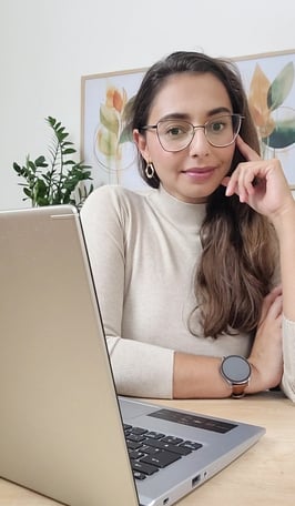 Mulher de óculos, suéter bege sentanda em uma mesa em frente a um notebook olhando para frente