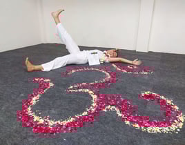 Eva lying on the ground next to a flower mandala, smiling happily while holding her Yoga Teacher Training diploma in Rishikes