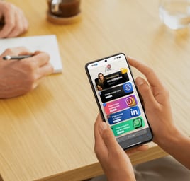 Pessoa segurando celular com mini site da Ajot Site aberto na tela, em ambiente de reunião sobre mes