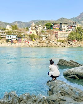 Eva standing by the sacred Ganges River in Rishikesh, India, with mountains visible in the background on a sunny day.