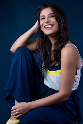mulher com cabelo ondulado, senta, sorrindo vestindo conjunto azul e branco em estúdio