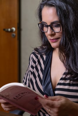 mulher com cabelos longos e escuros, de óculos, em retrato de perfil segurando um livro